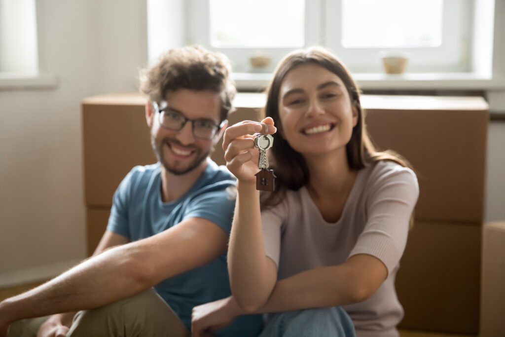 Happy young couple showing keys from hew home together, looking at camera, smiling, sitting at cardboard relocation boxes, feeling happy, enjoying moving into new flat, apartment, house